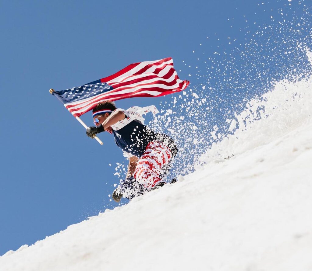 A skier on slushy snow carries an American flag, wearing USA themed pants, on a sunny day ski descent
