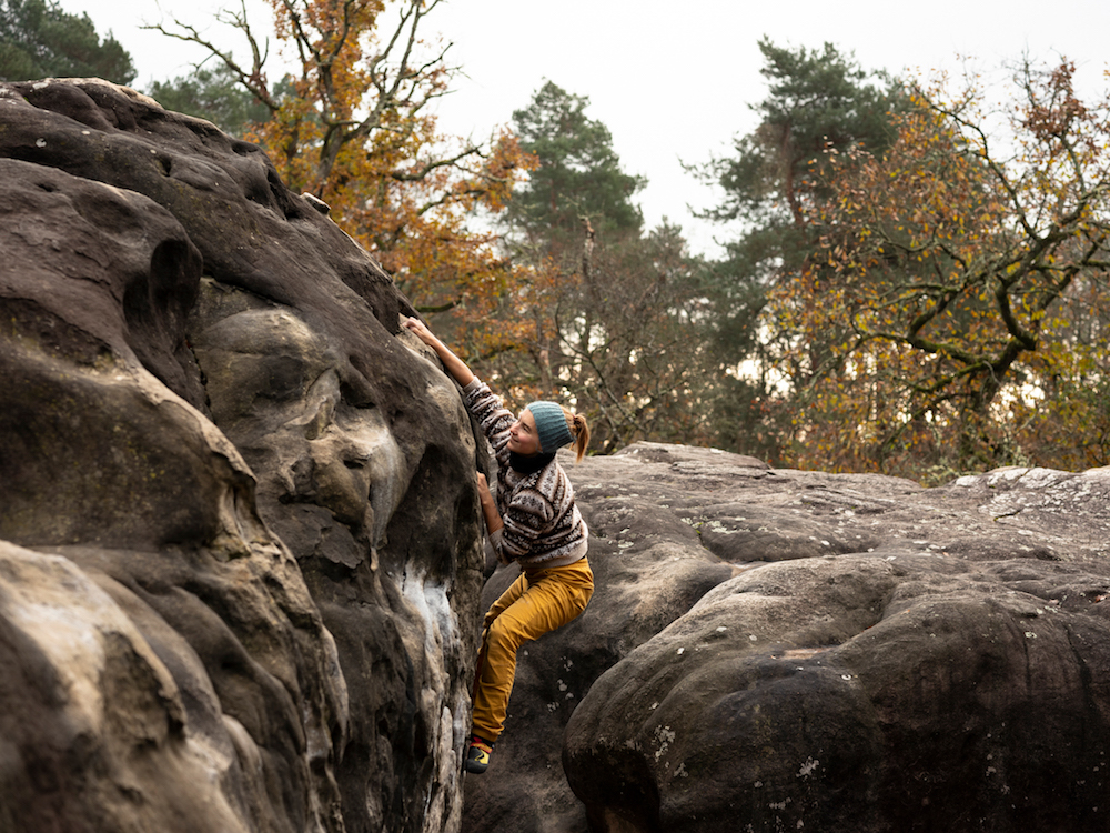 a person in yellow trousers is bouldering on smooth rocks, in front of yellow leaf trees suggesting its autumn