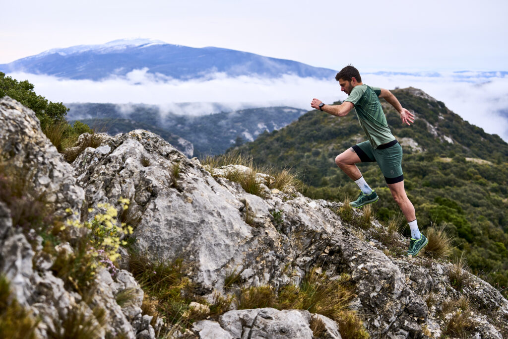 A man in green runs up seriously steep terrain, mountains and cloud in the background, uneven rock under his feet