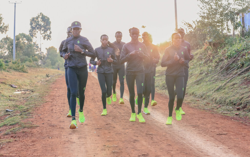 A team of Kenyan runners dressed in Scott black running kit and bright yellow trainers, running on a red-mud track