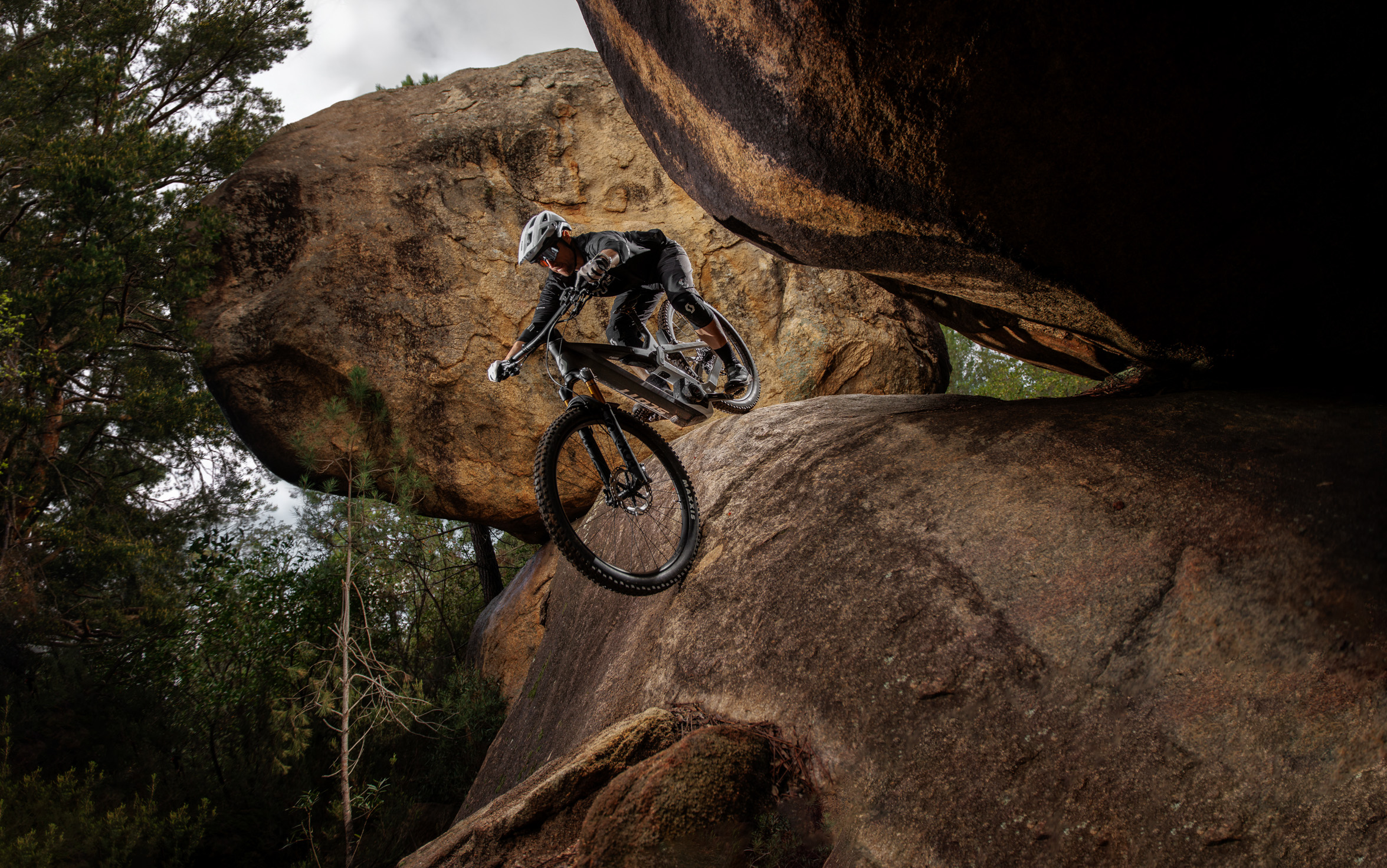 Mountain biker launches down a rock, two huge rounded rocks hanging over. It's an extreme image