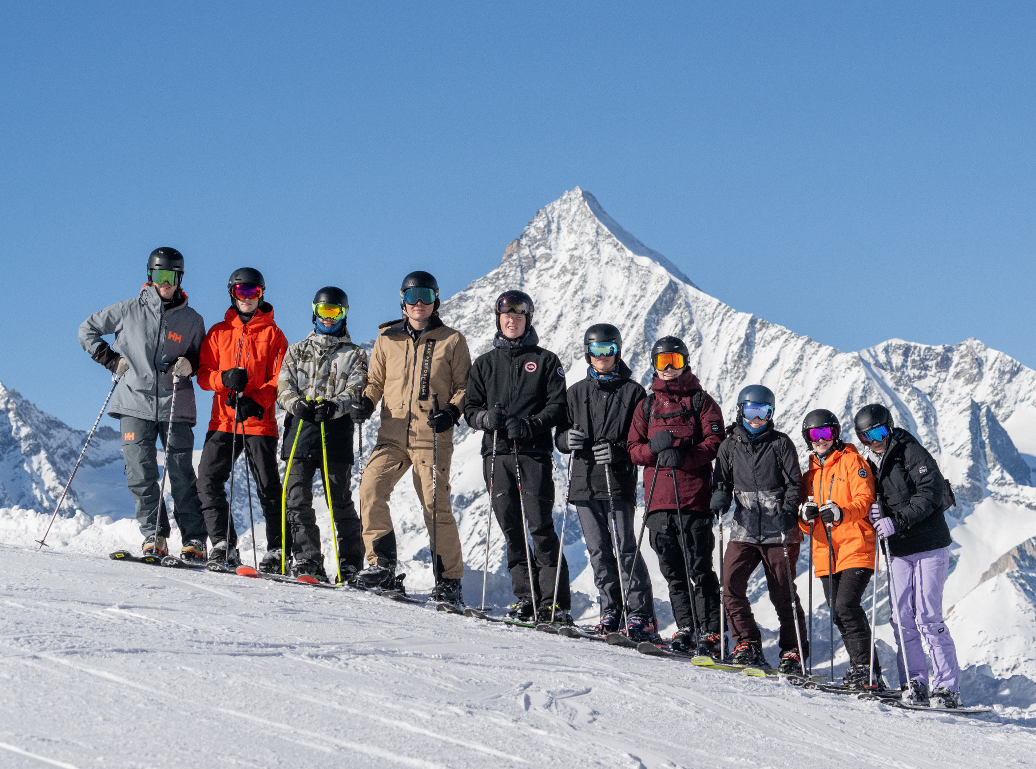 10 skiers line up along the edge of a piste, a high mountain in the backdrop, for a photo