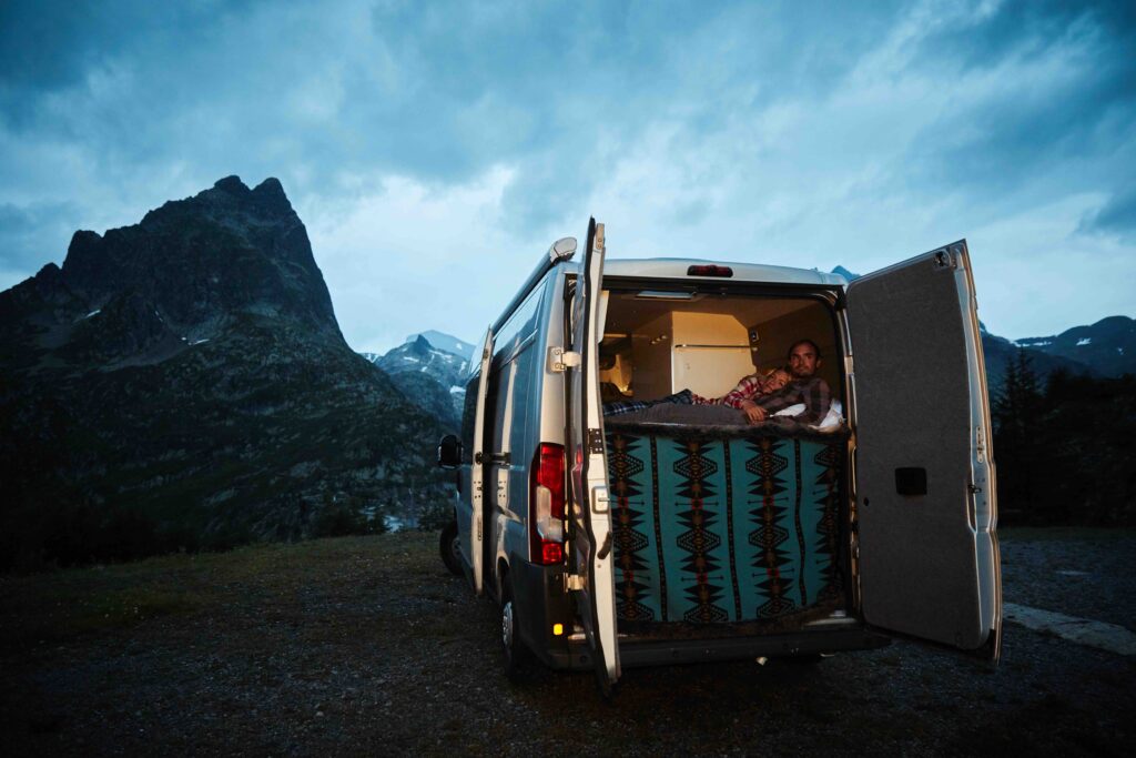 A couple snuggle in the back of a camper, back doors open looking out on the mountainous scene at dusk