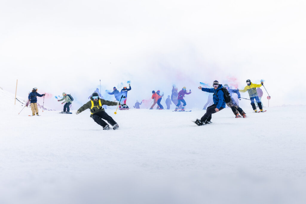 skiers and snowboarders mass ride down a gentle slope with coloured smokers