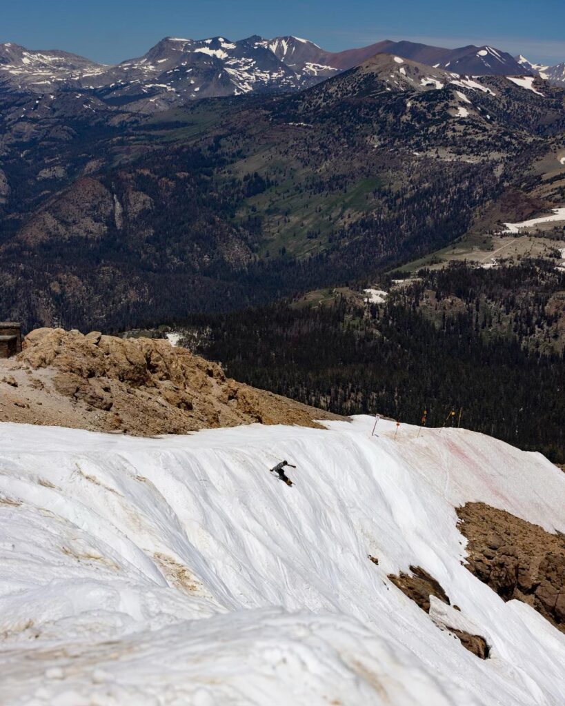 A solo skier heads down a steep, during what's clearly a summer ski session. Snow is textured and looks corny, mountains behind are nearly bare from snow, and rocks and dirt protrude through snow
