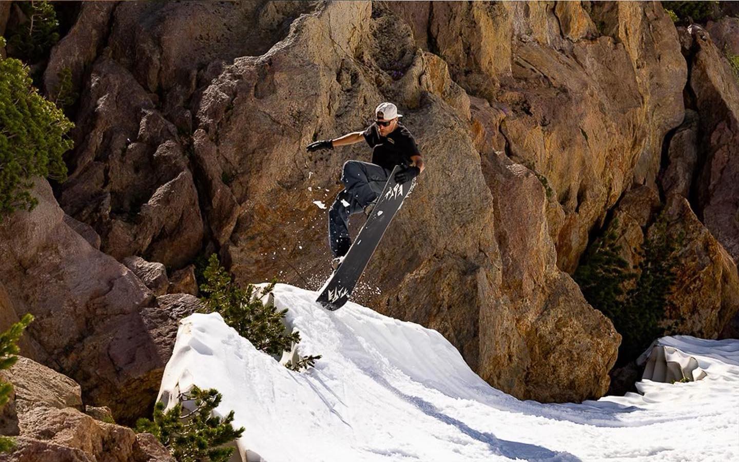 A snowboarder takes air on a side hit, grabbing nose. There's reddish coloured rock behind, with just a little snow to jump, and the rider wears a t shirt suggesting it's definitely spring summer skiing