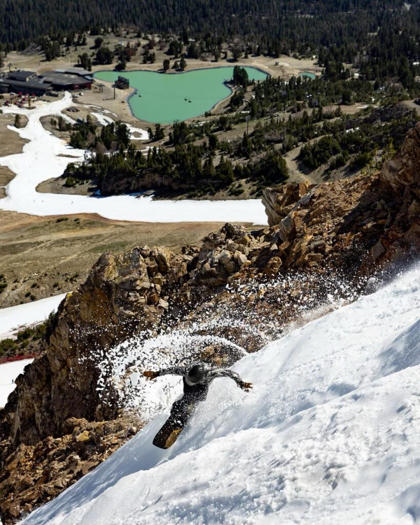 A snowboarder takes air on a turn on a steep slope with corn snow, the valley not far below looking very spring like with a strip of snow for a piste and a green summery alpine lake