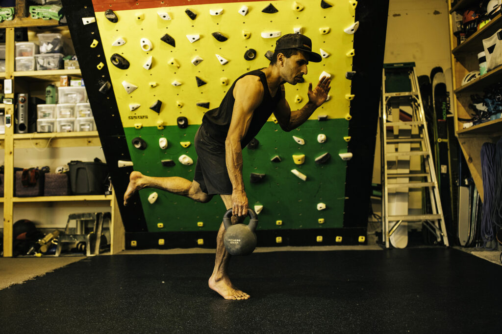 a man holds a kettlebell in one hand, standing on one leg, in front of a climbing wall