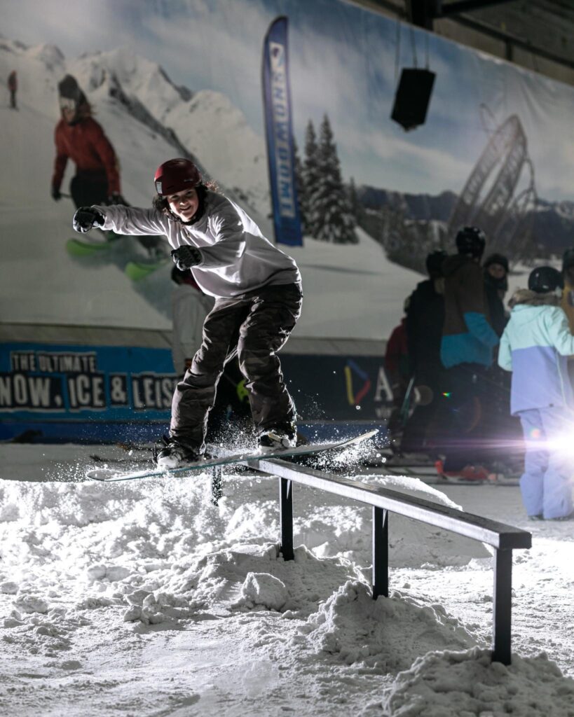 A snowboarder mid-rail in an indoor snow centre with a big smile on face