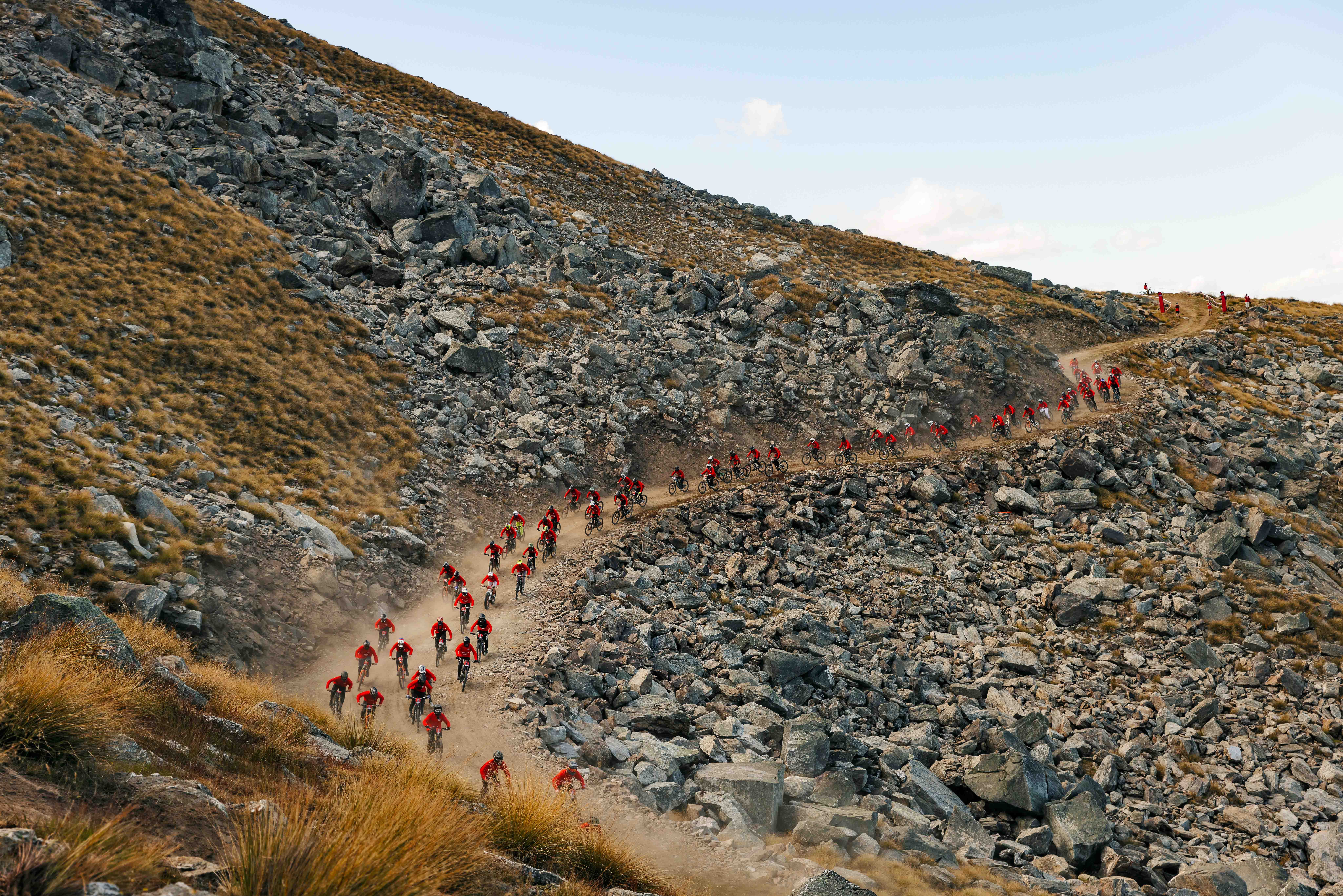 mountain bikers in red jerseys descend (en masse) down a red dusty rail, surrounded by rockfall and golden grasses, in the Cardrona Fox Hunt event