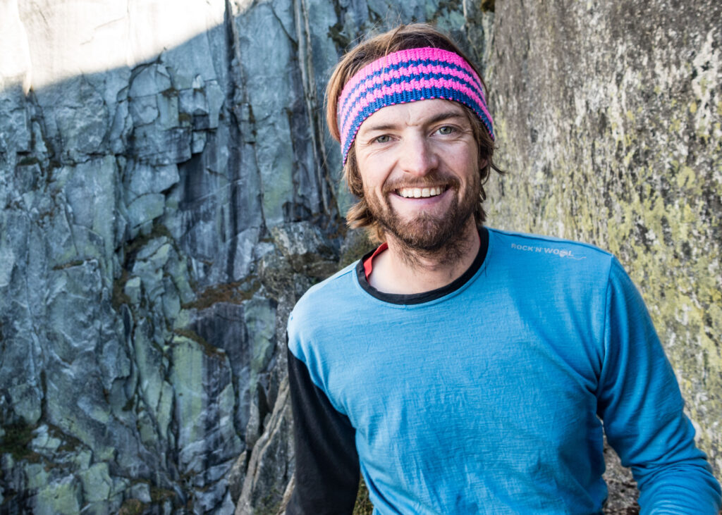 A man in blue with a pink knitted headband smiles to the camera, in front of a sheer rock race. This is Dan Louten, Swiss Mountain Guide talking pictured in Andermatt in summer