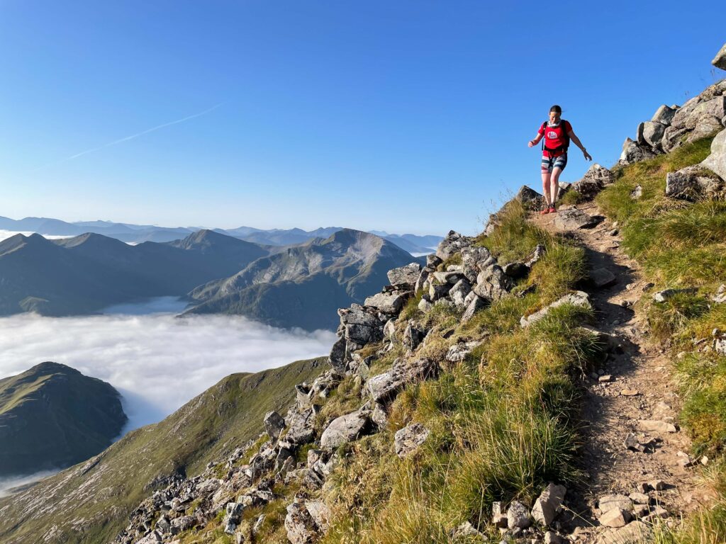 a rocky sky trail with a woman in a red vest running on a downward trajectory on mountain trail running trail, above the clouds high in the mountains