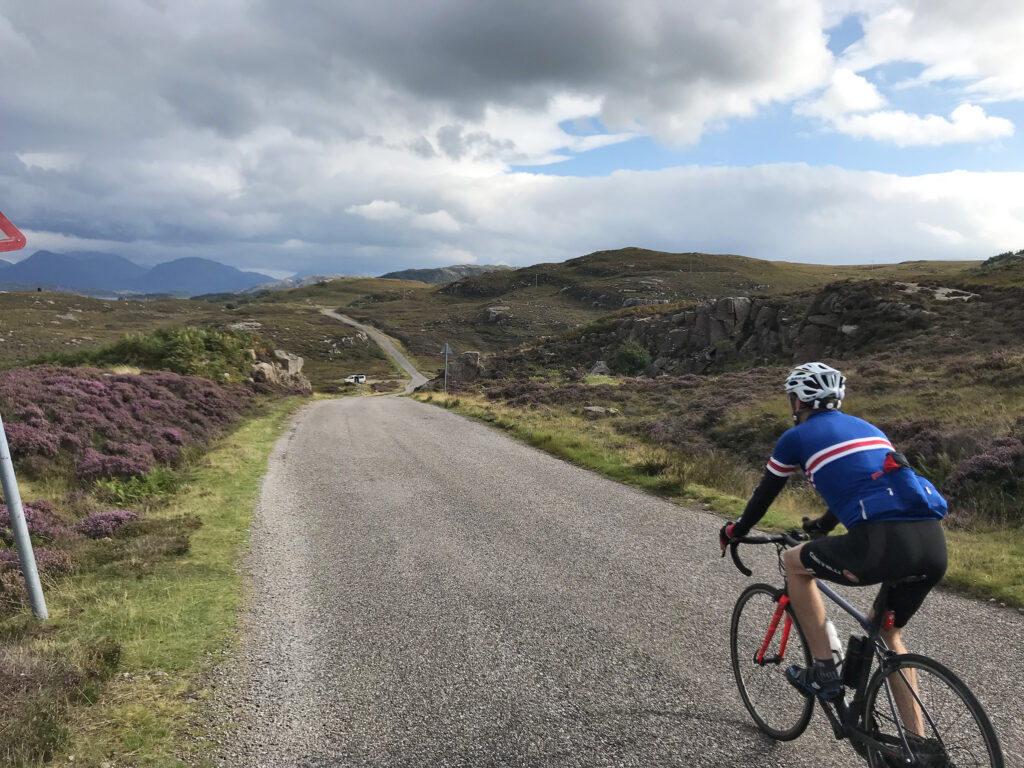 a road biker passes camera - back in shot - on a quiet paved road in the hills, pink heather on the side of the road, rolling hills in the distance