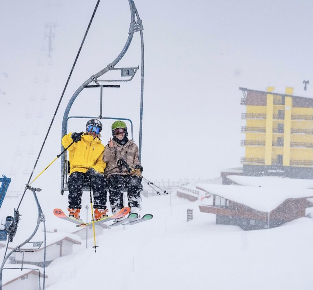 two skiers sit on a chairlift (bar up) waving and smiling as it snows around them