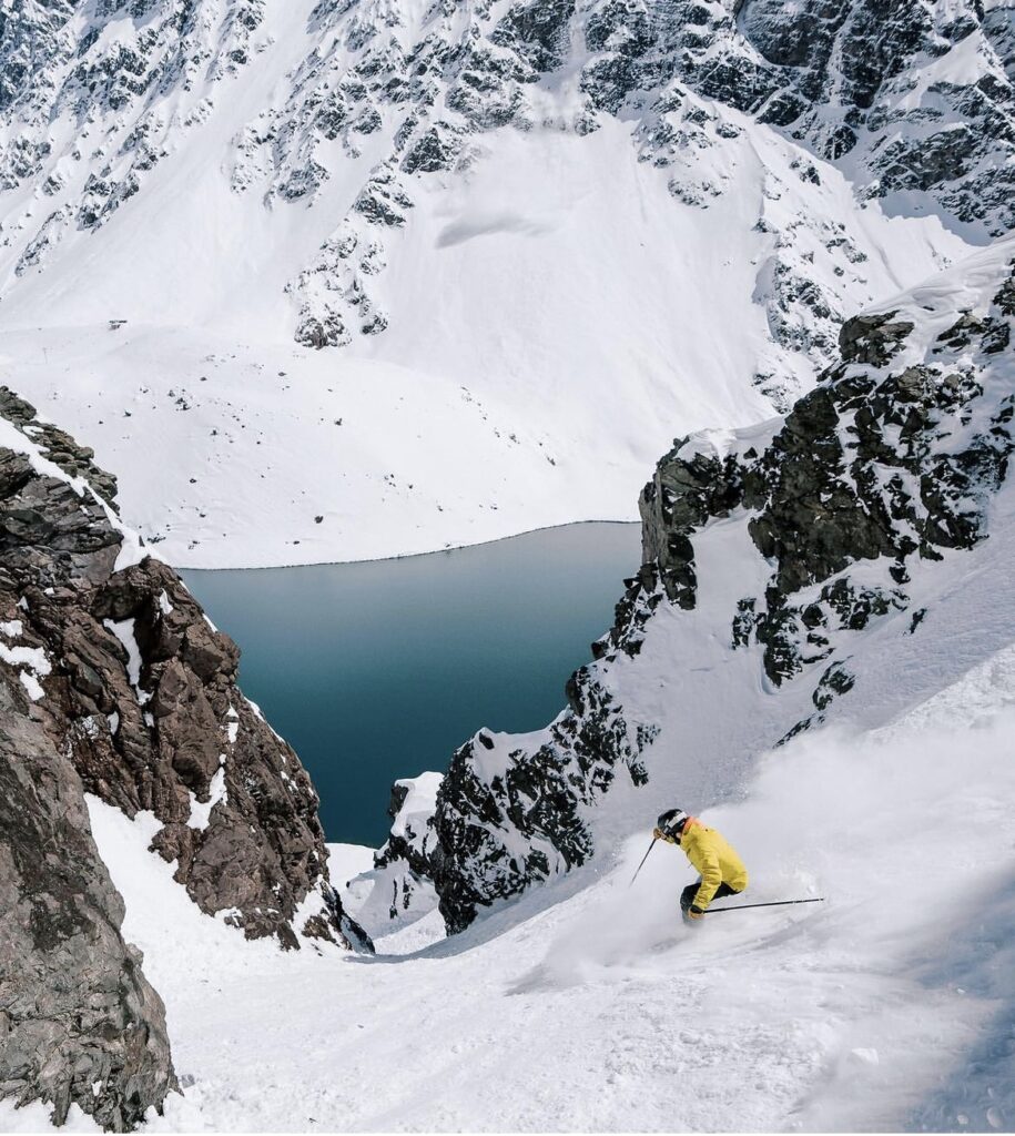 skier in yellow jacket skis in a couloir about to get narrow, above a blue lake