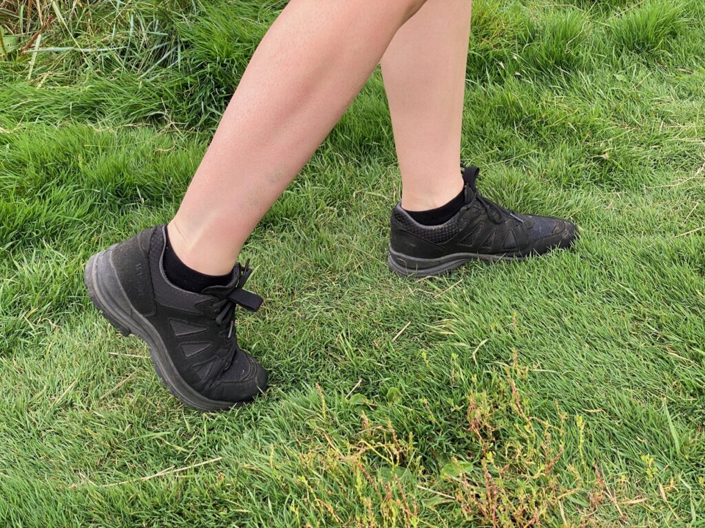 A black women's trail shoe (vegan it turns out) is shot in action on green grass, with bare legs