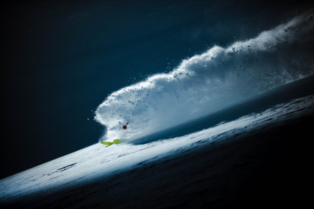 a shard of light illuminates a skier's powder cloud, darkness surrounding the area in an incredibly shot pro photo