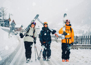 three skiers walk down a very snowy street, skis on shoulders, grinning big smiles