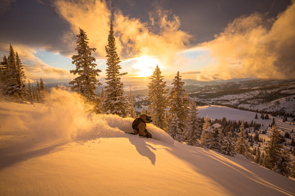 An orange light suggests sunset, with a skier making a turn in fresh deep beautiful powder, in the backcountry somewhere, tall fir trees as a backdrop and low lying valley looks like USA