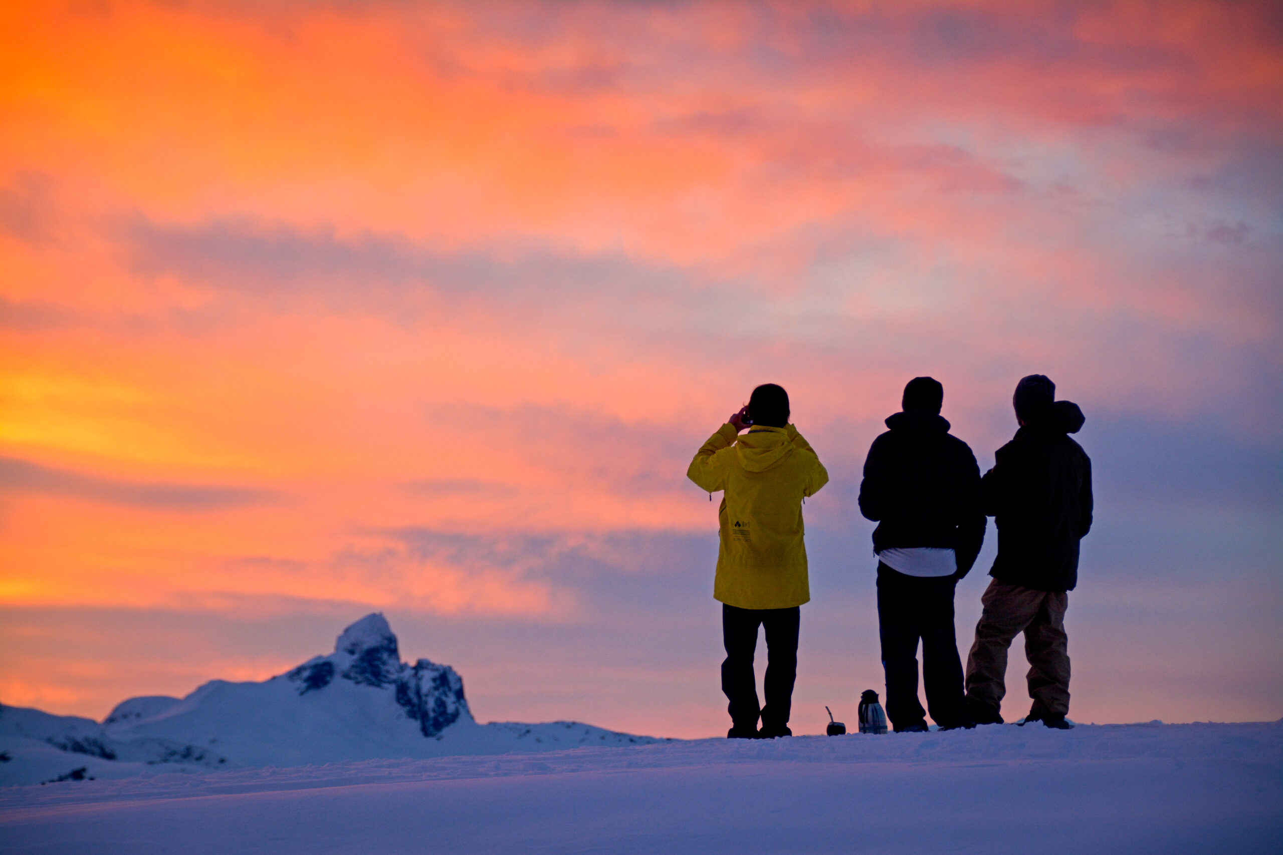 three backcountry skiers stand atop a mountain looking out to Blackcomb peak in the distance during a pink-cloud-sky sunset