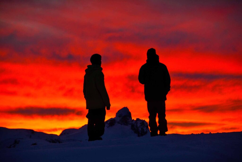 silhouettes of two people standing on the top of a mountain, the sky burning red behind at the last moment of dusk