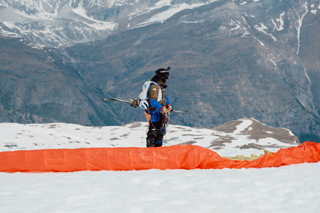 A skier stands strapped up to parapente, skis attached to backpack, witht eh kite on the floor, on the mountain side. He looks about to set off