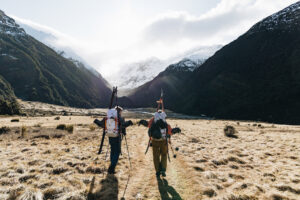 two skiers, skis on backpacks, walk through a grassy valley, snowy mountains ahead and above