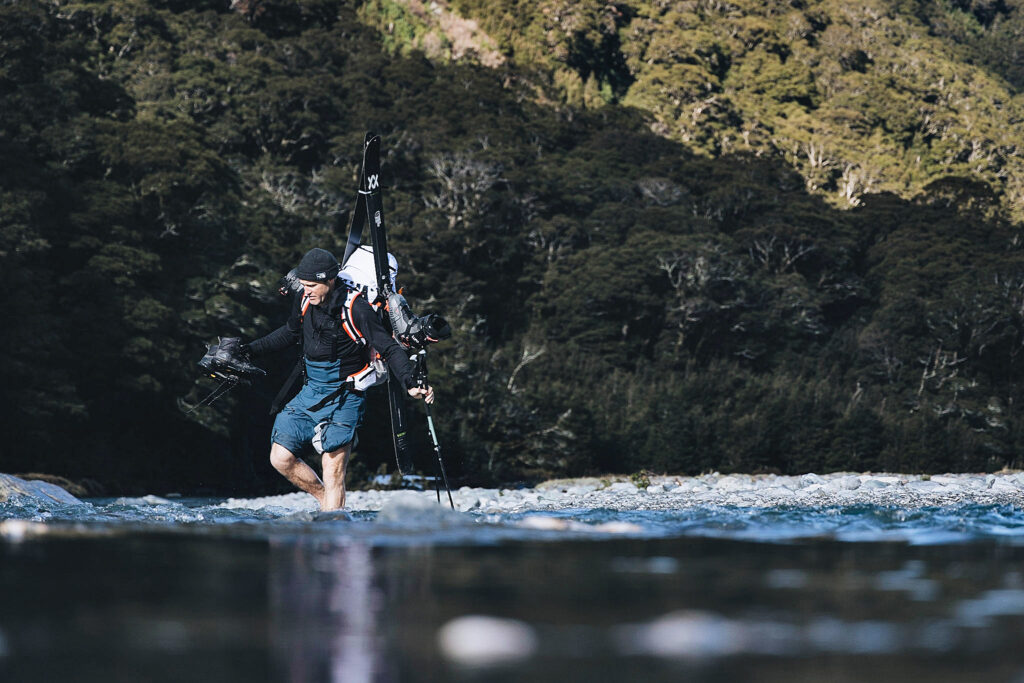 Skier Sam Smoothy walks, skis on back, wades through a river, trousers rolled up