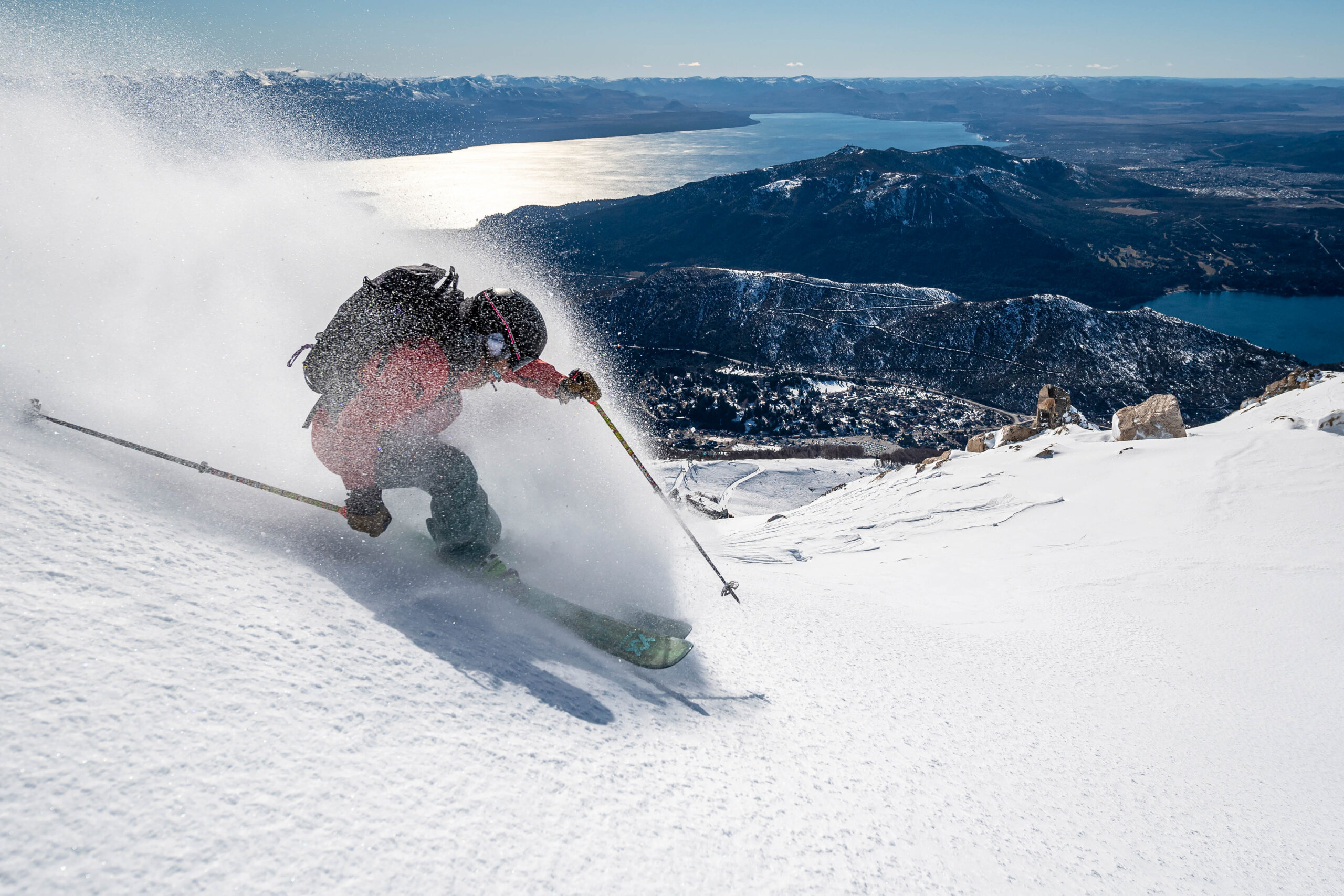 A snowless valley and lake is visible in the distance, whilst a skier (Ingrid Backstrom) makes a turn in light virgin powder