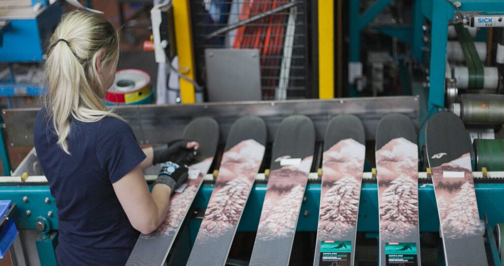 skis lined up in a factory, a woman with a pony tail tending to them