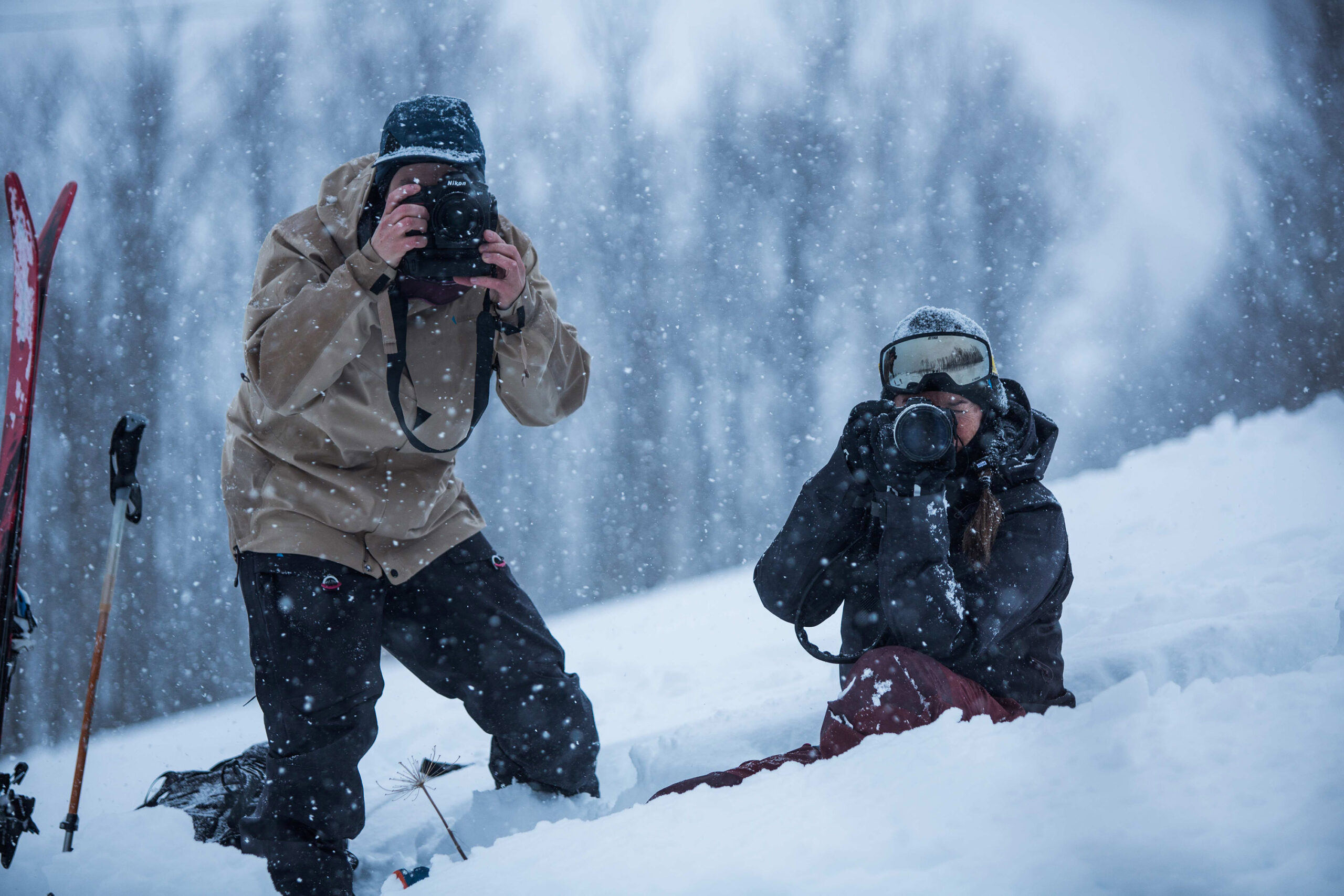 two photographers look through the lens of their camera, right at the picture-taking camera, standing and kneeling in deep snow, the skeletons of trees blurred in background