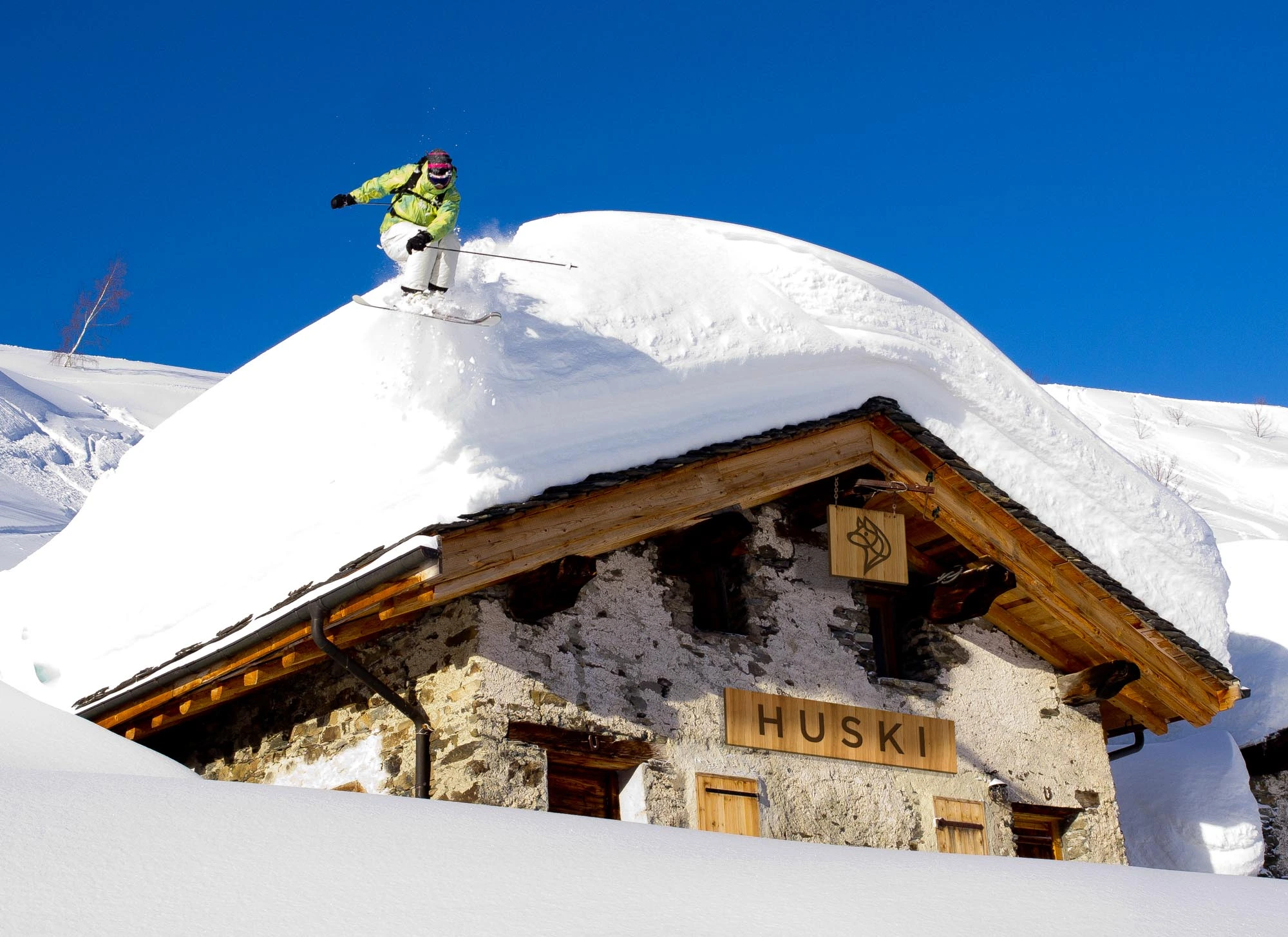 a stone chalet covered in snow, with a skier jumping off the roof maybe 2 metres deep with fresh snow