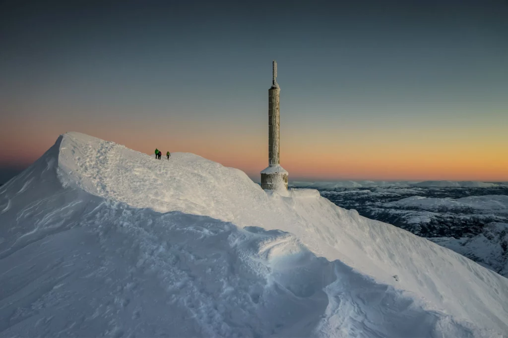 Top of Gaustatoppen, Norway, with a tall communications tower, pictured at dusk, with three people walking the ridgeline