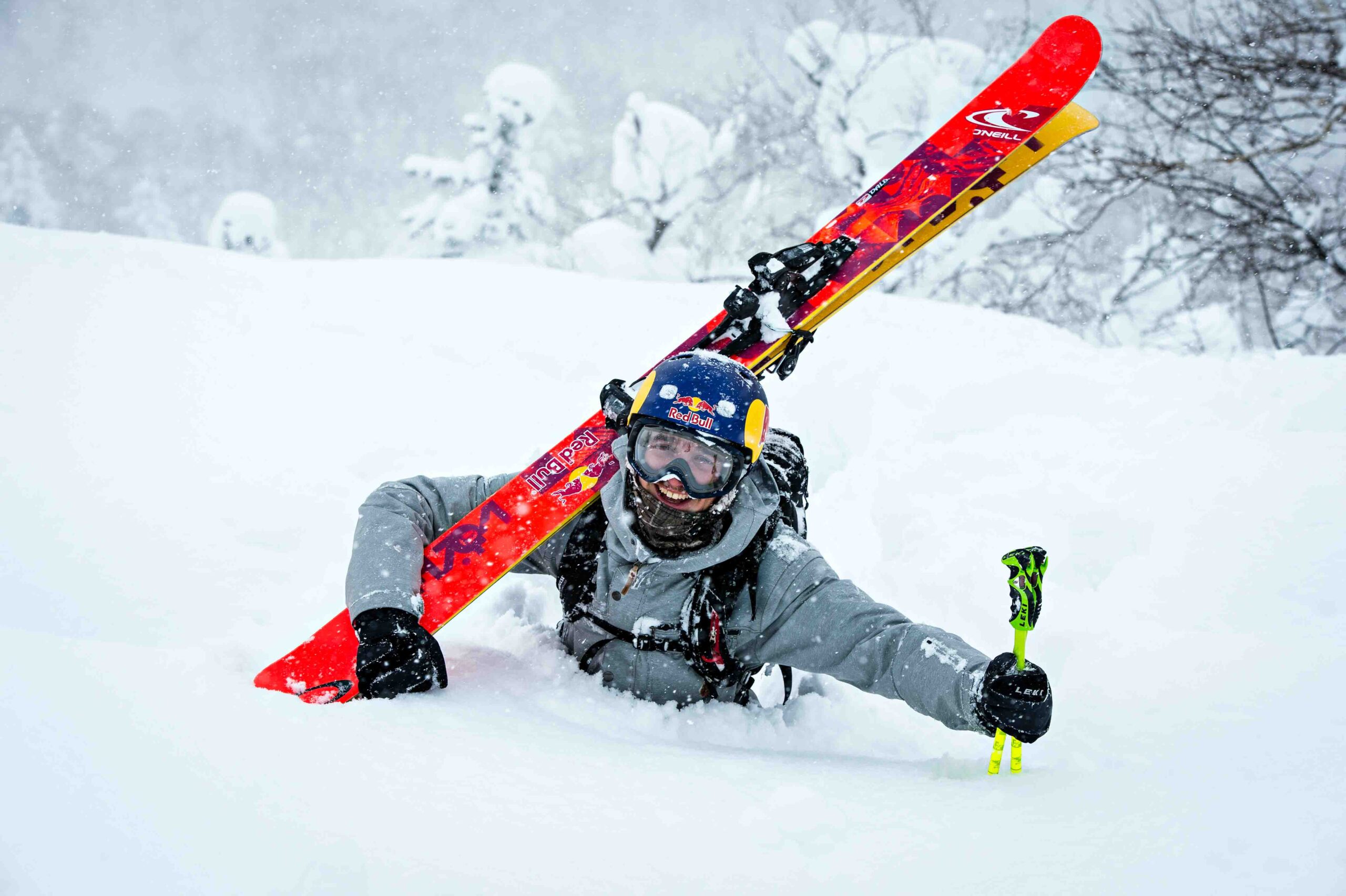 a skier wades chest deep through snow, orange skis carried on shoulder