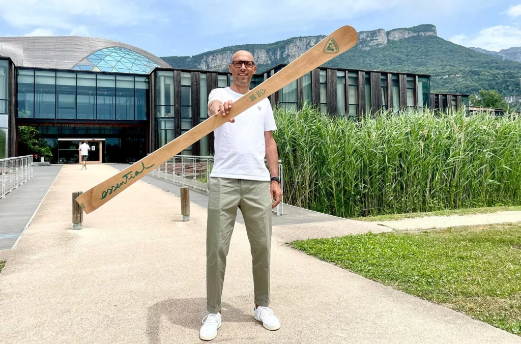 man holds the Rossignol Essential ski in hand towards the camera, outside the factory, a green-covered mountain in the background