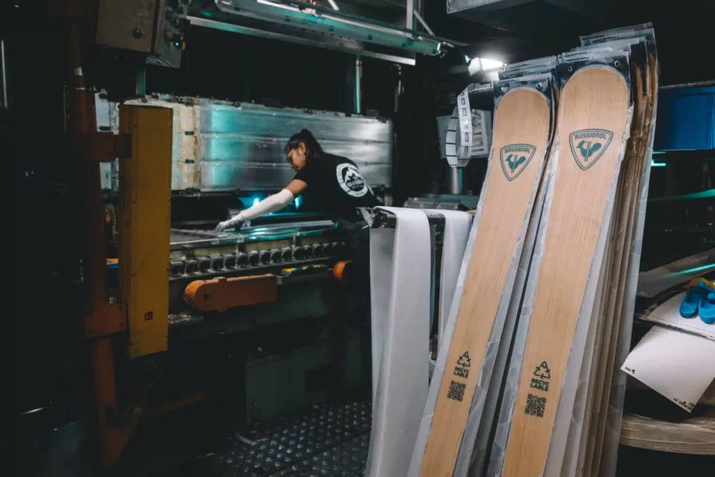 The wood-look-finish of the Rossignol Essential recyclable skis are stacked up in the factory, a woman working on a machine behind