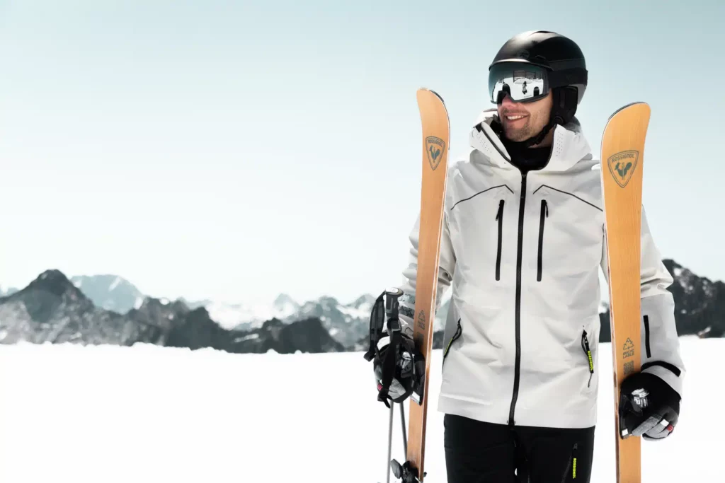 man holds recyclable wood-finish skis from Rossignol in each hand by the binding, the top-of-mountain scenery behind blurred