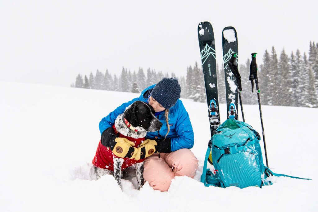 a woman kisses her dog out in the countryside, with with skis and poles and pack suggesting they're out touring -  in a competition post to win Baïst gloves