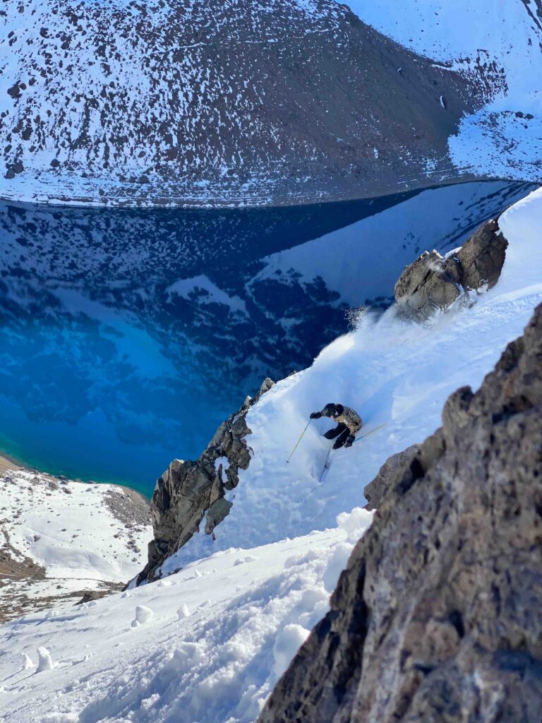 Freeskier Ingrid Backstrom skis a couloir, above a lake reflecting the mountains above it, out of shot of the photo