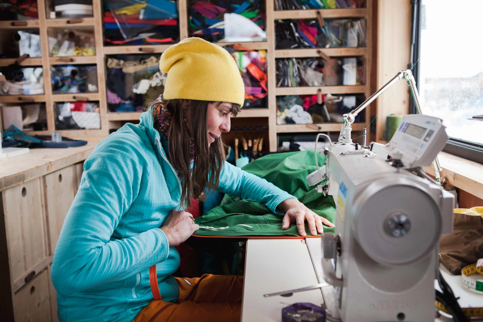 a woman in yellow hat sits at a sewing machine repairing a green jacket