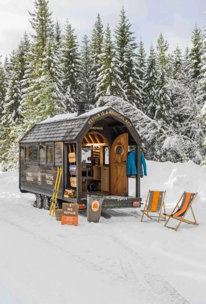 Scandi style wood Worn Wear hut-van by Patagonia, with deck chairs outside, a jacket hung up and yellow skis resting against it, parked on the snow in front of snowy fir trees
