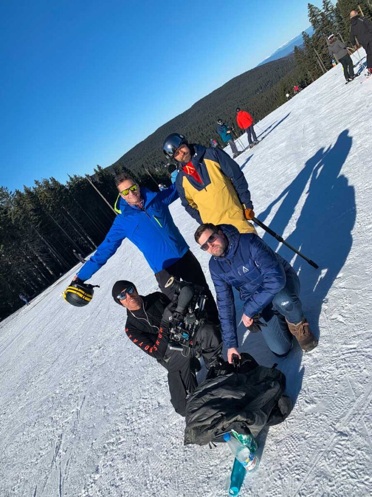 four men on snow, one a camera man holding a huge cam