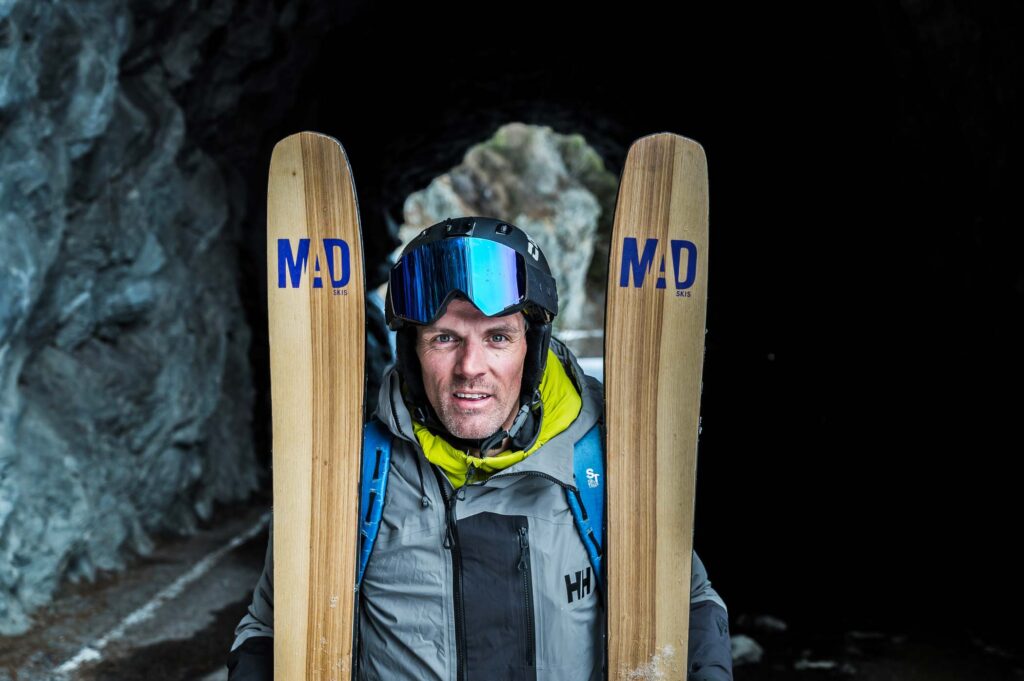 man smiles into camera in a rock tunnel, holding skis tips up, either side of his face