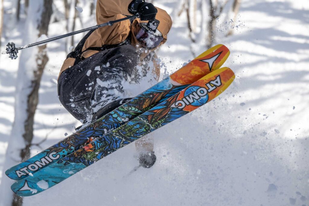 skier tucks, jumping sideways, in the trees off piste, the colourful bases of his skis the image focus