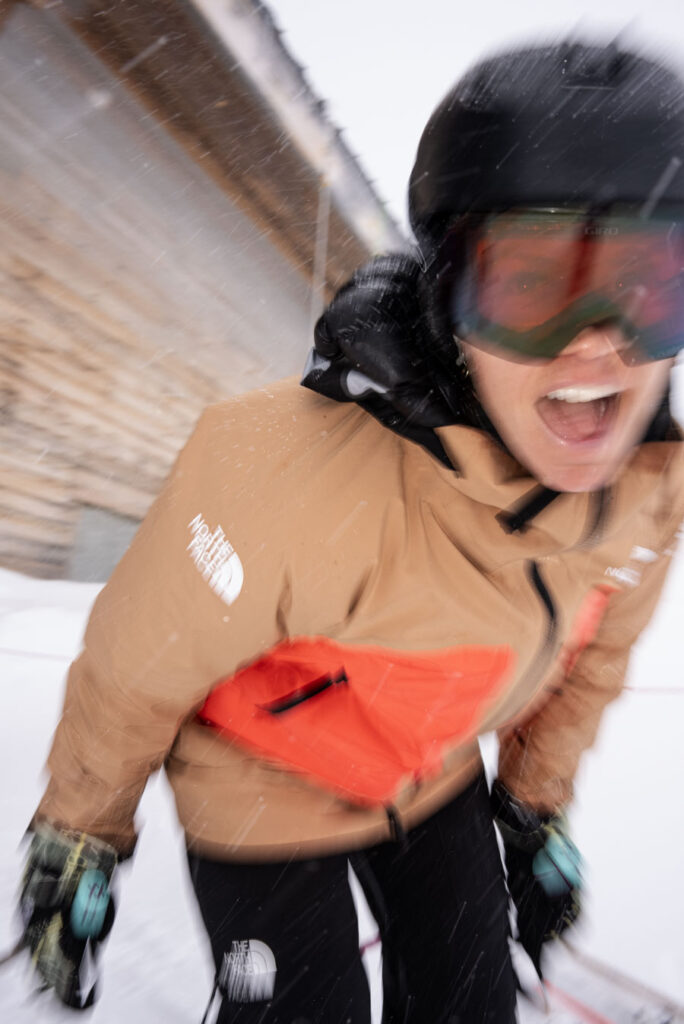 blurry figure of skier comes at camera, mouth open in celebration, by a wooden chalet on a snowy day