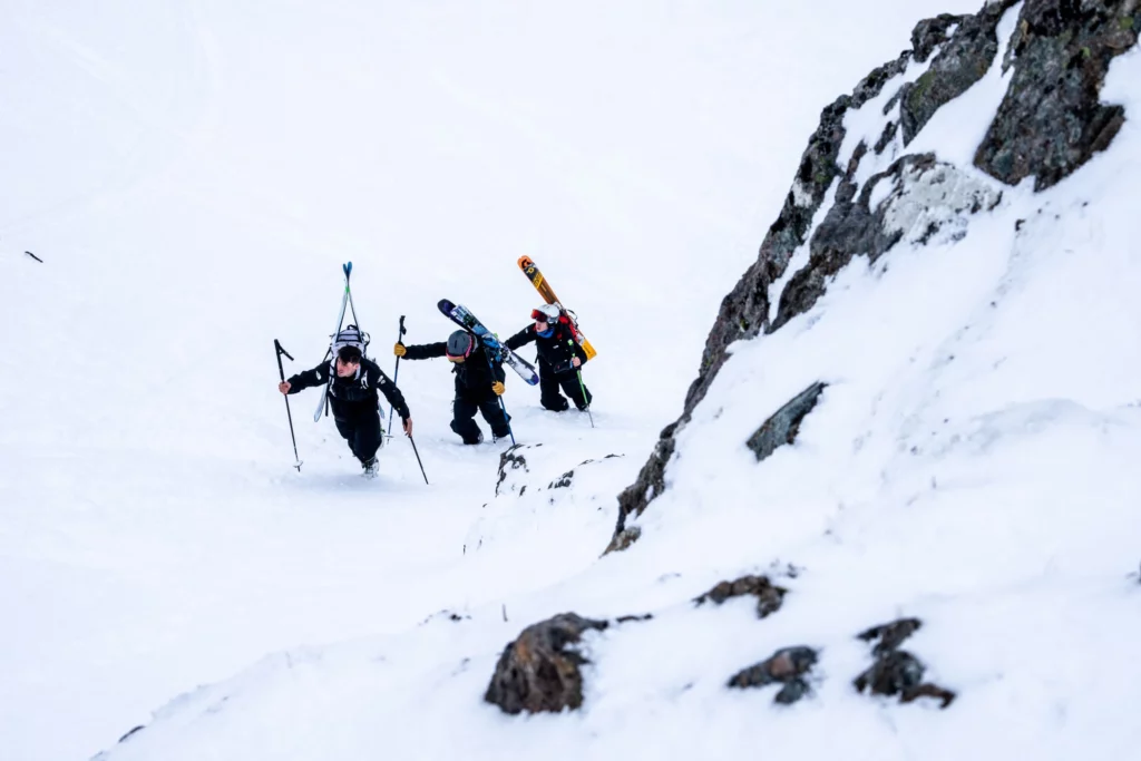 three skiers boot up a steep hill, skis on packs, next to a rock band