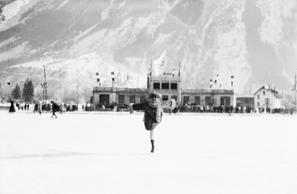a black and white image of a woman ice skating, gliding on one leg towards the camera, with snowy mountains blurred behind, a photo from the first Winter Games in Chamonix