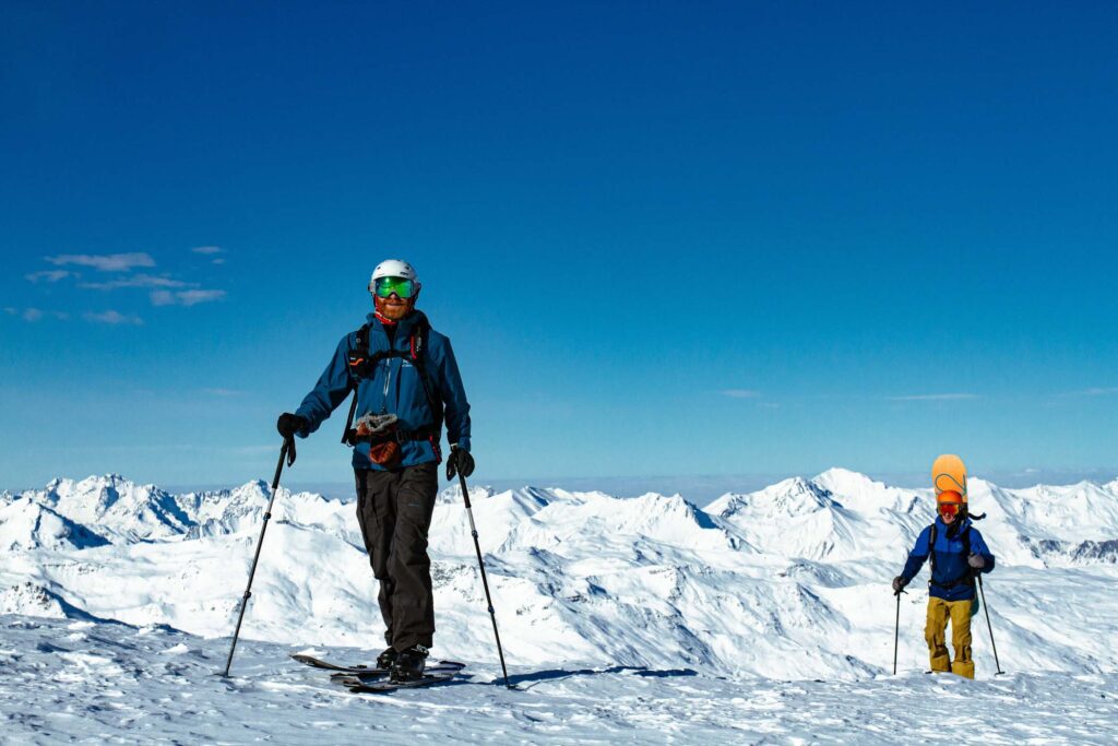 a splitboarder and snowboarder walk uphill at the very top of a mountain with a huge snowy Three Valleys vista behind