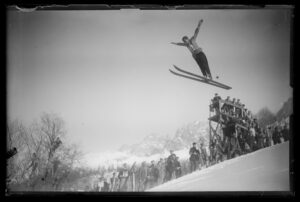 a skier, arms wide, jumps big in a black and white Olympics photo from 1924, the spectators on a wooden frame and lining the slope beside the jumper
