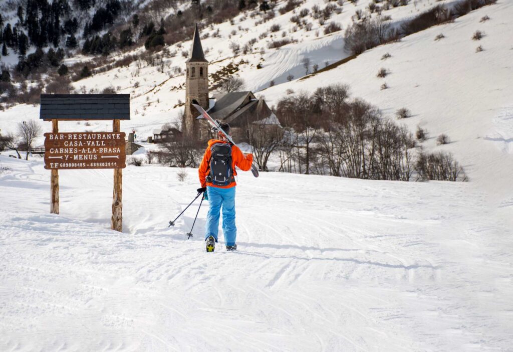 a skier in blue and orange walks away from the camera, skis on shoulder, towards a mountain church surrounded by snow